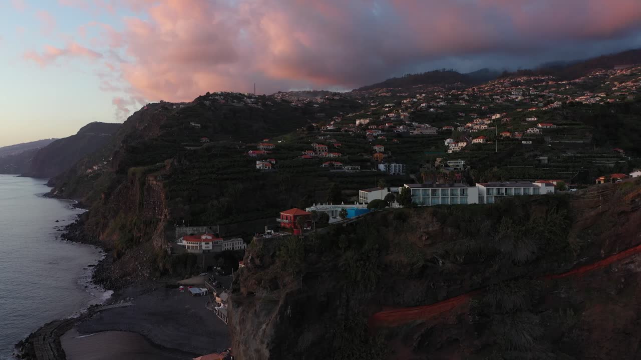 Aerial shot moving forwards and tilting up, focusing on the cliff of Ponta do Sol on Madeira island. Dramatic and colorful storm clouds in the background during the sunset on the coast.
