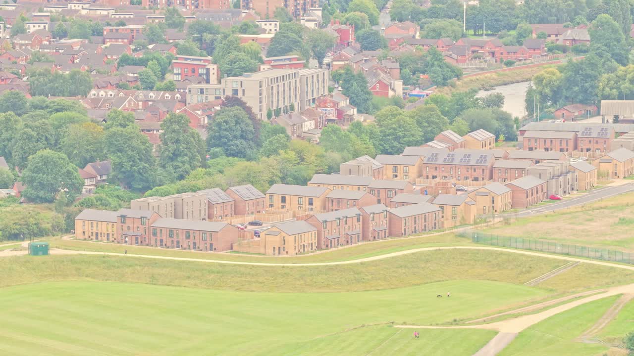 Rows of contemporary brick townhouses and apartments stand near the River Irwell alongside open green space in Salford, England, captured in static aerial shot