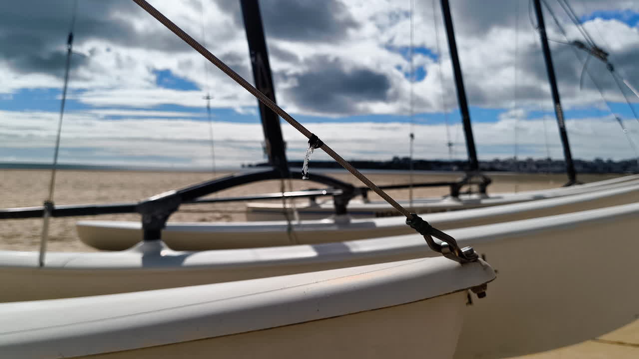Close-up view of sailboat rigging on a sandy beach under a cloudy sky, with distant shoreline and calm waters visible in the background.
A detailed view of sailboat ropes and masts on a quiet beach.