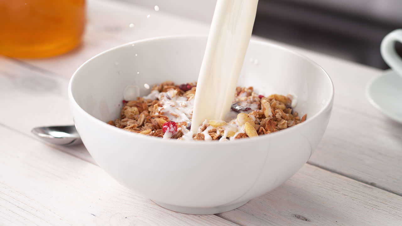 preparar un desayuno saludable: verter leche en un cuenco de cereales de granola crujientes colocados en una mesa de la cocina, en cámara lenta