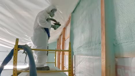 man in disposable suit and respirator spraying foam insulation onto an exterior wall of a new construction while standing on a scaffold