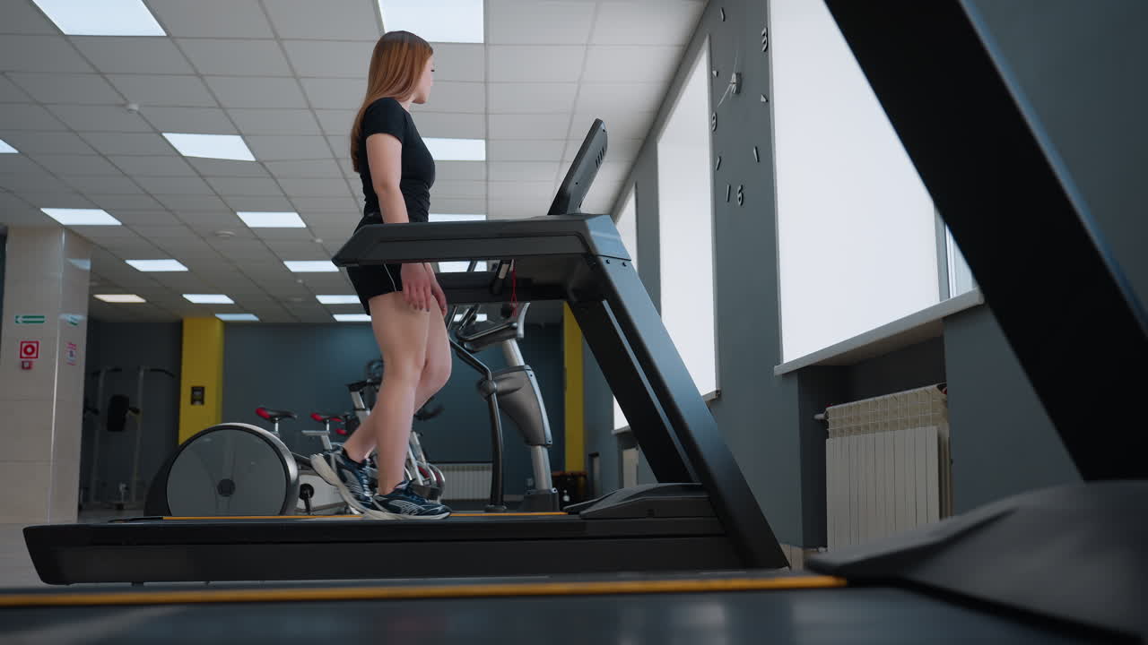 close up of treadmill machine under digital console with lady walking on belt next to pillar with red sign in bright modern gym studio featuring tiled floor and exercise bikes