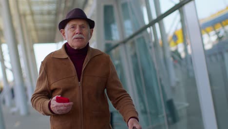 Abuelo-Turista-Senior-Hombre-Caminando-En-El-Vestíbulo-Del-Aeropuerto-Internacional,-Usando-Teléfono-Móvil,-Enviando-Mensajes-De-Texto