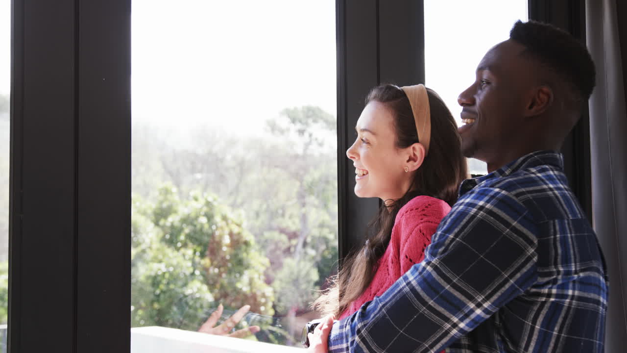 Happy diverse couple looking out window, smiling and embracing, slow motion, copy space