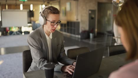 A-confident-blonde-girl-in-white-headphones-and-a-business-suit-sits-at-a-table-in-the-office-and-types-and-works-on-a-laptop.-A-middle-aged-woman-with-glasses-in-a-business-uniform-works-at-a-laptop-in-a-modern-office