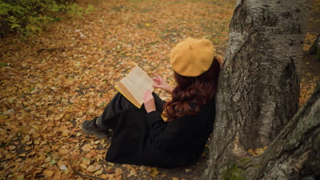 overhead view of woman seated outdoors in autumn forest leaning against tree, reading a book, wearing yellow beret and black coat, she glides her hand along book's edge