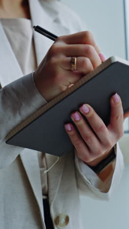 woman in light jacket standing at the big window. lady holding a notebook, writing something down there and closing it. close up. vertical video