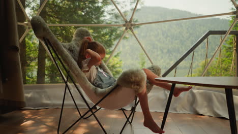 little girl basking in cool fur chair and getting comfortable for afternoon nap. child watches landscapes of forest and mountains behind transparent window of glamping