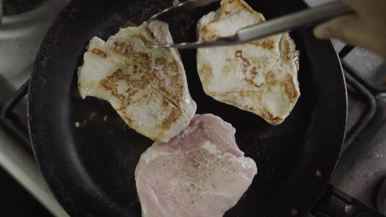 Primo piano ravvicinato dall'alto di bistecche di lonza di maiale crude girate con pinze di metallo in una padella calda, che mostra il processo di doratura e rosolatura.