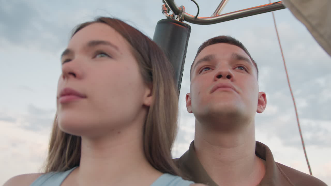 young man stands close to sister in hot air balloon hold basket edge gaze upward at burner flame erupting against pastel sunset sky while siblings share adventurous serene