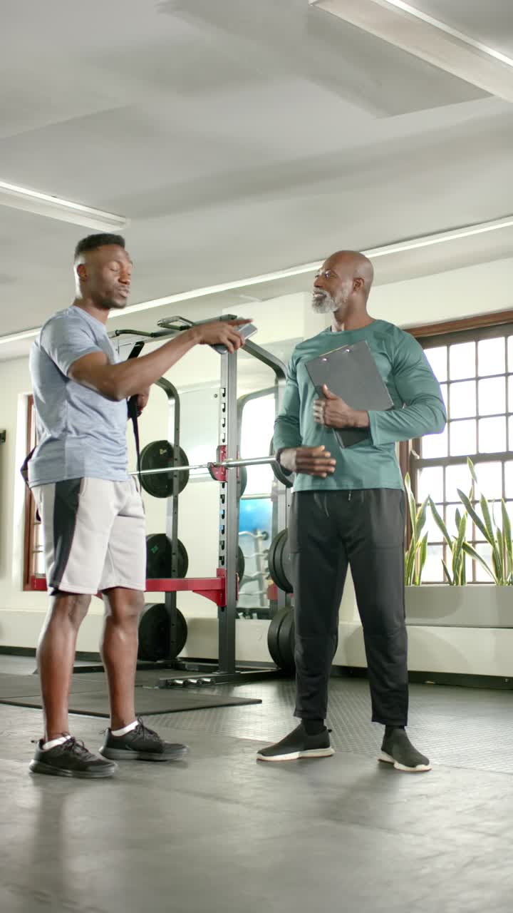 Vertical video: African American trainer pointing squat rack, outlining workout and trainee nodding