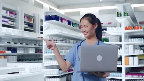 doctora asiática con estetoscopio sosteniendo una computadora portátil, apuntando a un lado y sonriendo en la farmacia