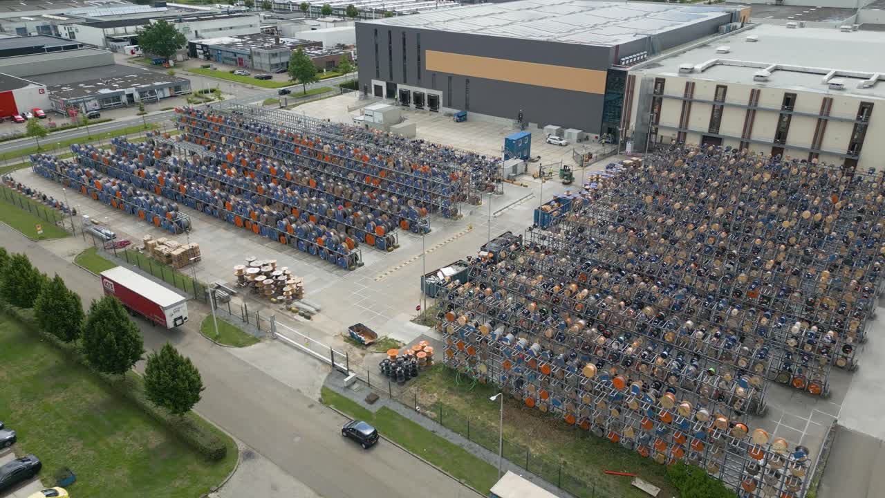 A wide drone shot showing an industrial outdoor storage yard filled with large stacks of metal racks containing cables, electrical components, and wiring supplies