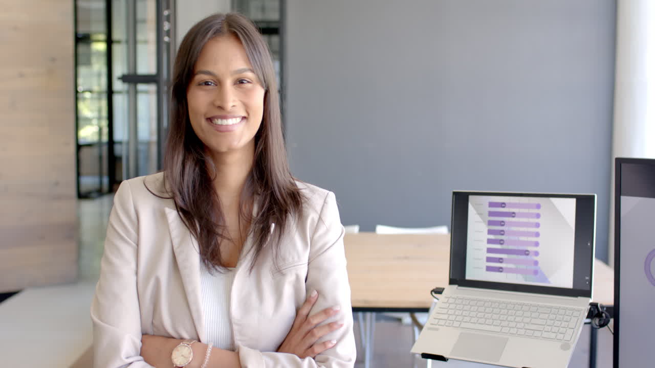 Smiling businesswoman standing with arms crossed next to laptop in office