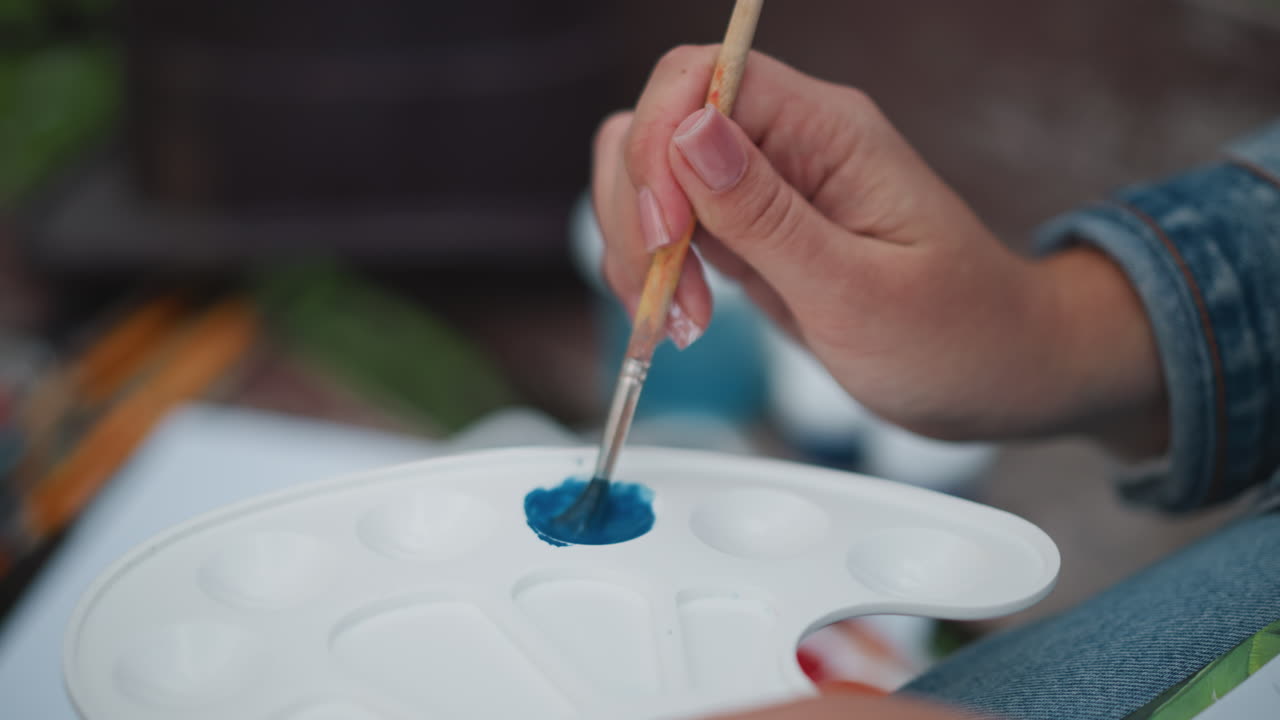 パレットで青い絵の具を混ぜる手のクローズアップ。デニムの袖をまくった人が優しく筆に絵の具を乗せている。プラスチックパレットのくぼみには鮮やかな顔料。背景にはぼかされた瓶と筆が見え、穏やかな屋外での創作活動の様子。