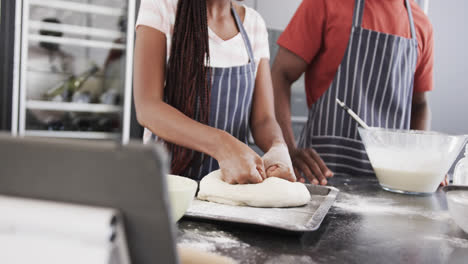 happy african american couple in aprons baking using tablet, kneading dough in kitchen, slow motion