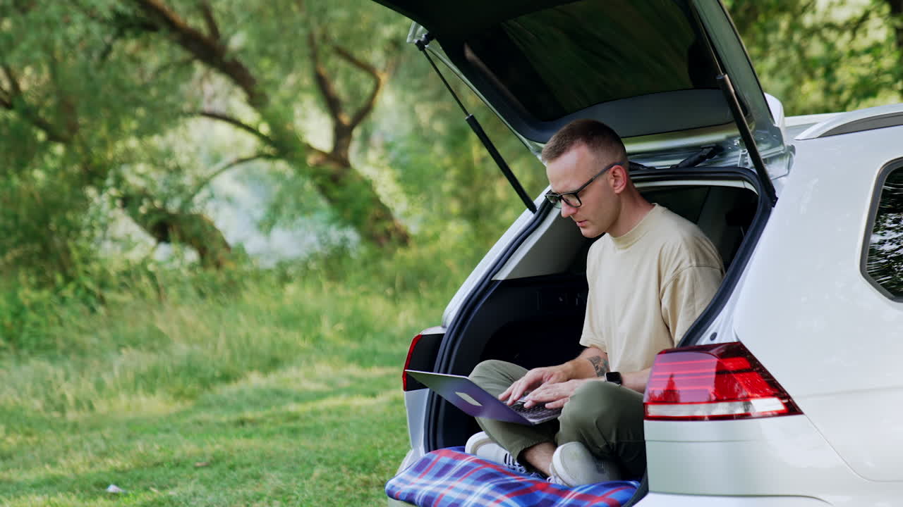 Freelancing man works on his laptop. Entrepreneur works outdoors sitting in his car trunk.
