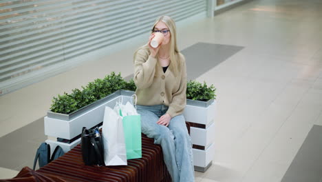 young woman sitting on bench in well-lit mall with shopping bags nearby, enjoying a peaceful moment, relaxing after a shopping spree in modern indoor setting with stylish atmosphere