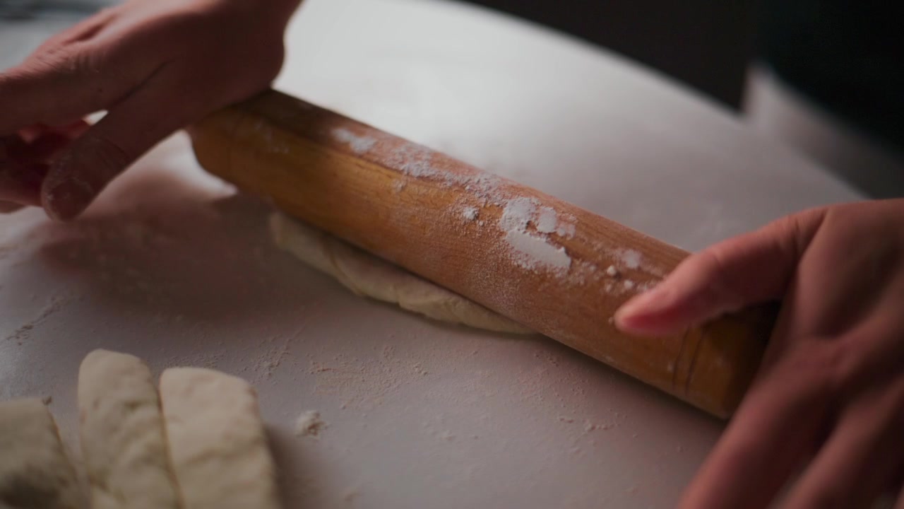 Close-up of hands rolling dough with wooden roller. Flour dusts surface as dough flattens. Person focuses carefully on even thickness. Perfect for cooking, bakery, or food preparation content