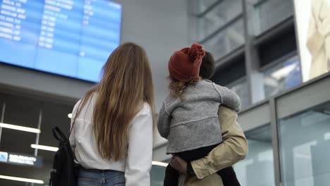 unrecognizable young family stop against and look to flight schedule board. departure area, midfield concourse of modern