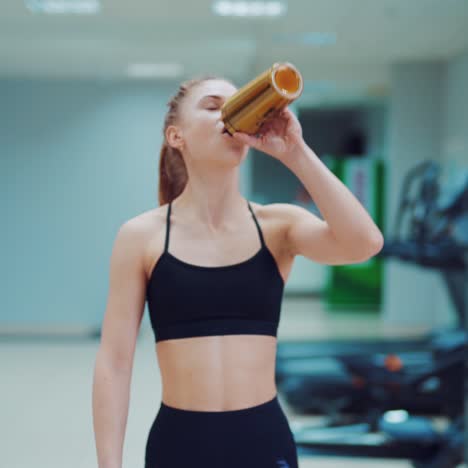 a young woman in black sportswear walks through the gym with a shaker in her hands and drinks water from it. fitness club. blurred background