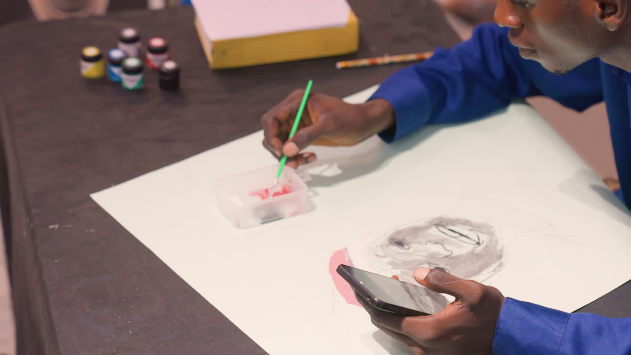 Black man painting on white paper, focused artist mixing black and gray paint, brushwork detail, smartphone reference held in hand, small paint pots and palette on tabletop, warm studio lighting
