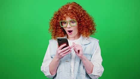 concepto de victoria en línea. joven mujer pelirroja rizada con gafas navegando por la web en el teléfono inteligente, leyendo grandes noticias y saltando