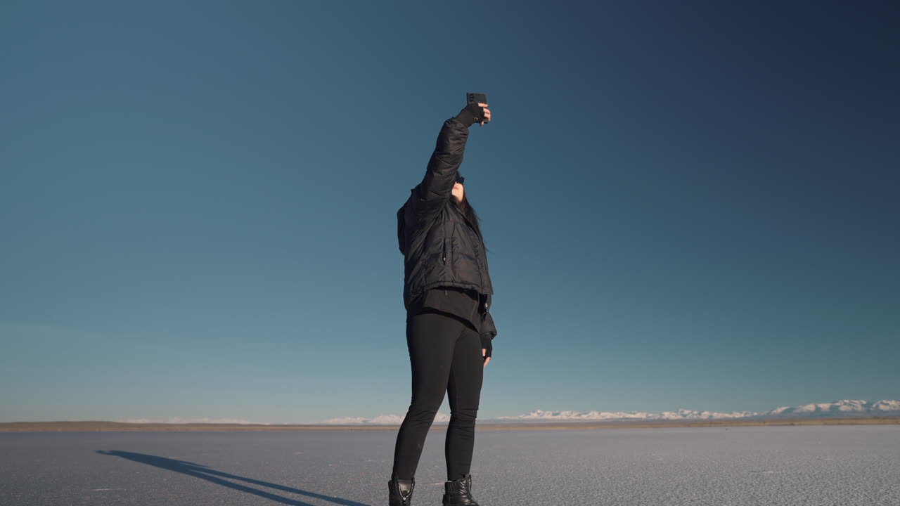 Giovane donna in abiti invernali alla ricerca del segnale mobile in un vasto deserto di distese saline sotto un cielo azzurro.