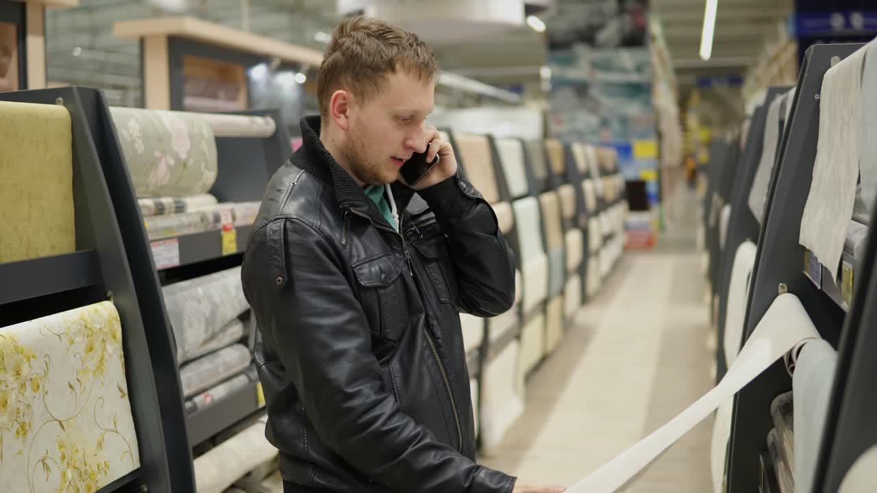 Young man is calling someone to be consulted while choosing wallpapers in supermarket. He is looking at the wallpaper, touching it and thinking which one is better