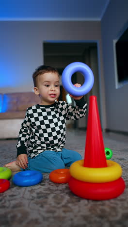 lovely two-year old toddler sitting on the floor building a pyramid. cheerful baby boy focused on his game at home. vertical video.