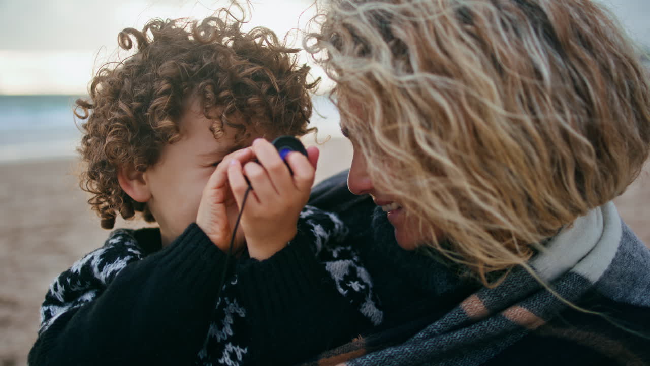 Bambino in primo piano che guarda con un cannocchiale sulla riva dell'oceano. Famiglia che ride e si rilassa all'aperto.