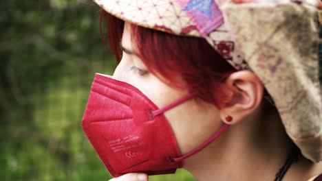 Side-view-of-a-red-haired-girl-covering-her-face-with-a-medical-ffp2-mask-in-a-green-outdoor-environment