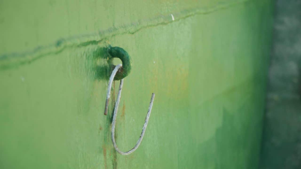 Gros plan sur une coque verte, corde effilochée, ligne d'amarrage laissée après le quart de travail d'un docker, surface métallique patinée, peinture écaillée et petit anneau de rouille, subtile saleté côtière et palette de couleurs sourdes, faible profondeur de champ.