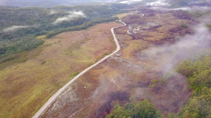 Volo aereo su montagne nebbiose e foreste verdi e rosse in Tasmania, Australia, con inquadratura grandangolare estrema a distanza, in movimento in avanti e inclinazione verso il basso.