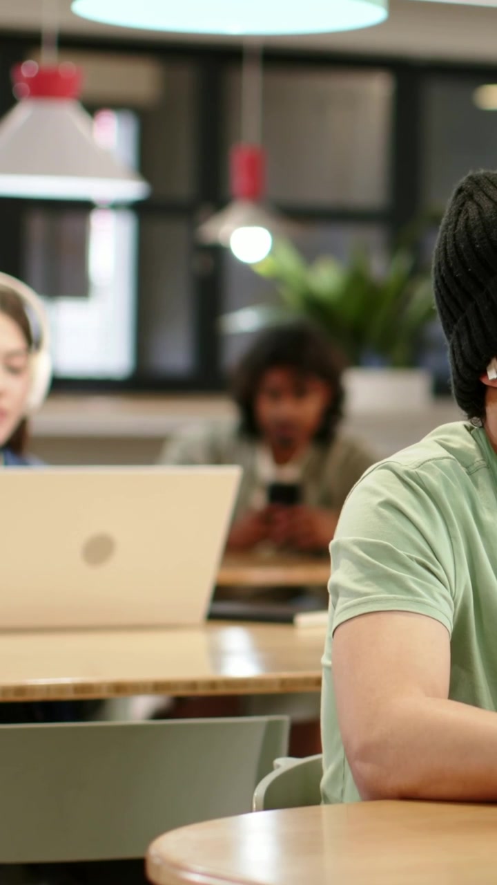 Vertical video: Adjusting earbud after slip, beanie man in green tee at study table, copy space