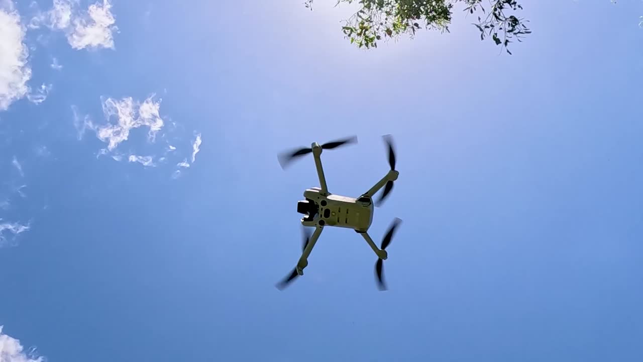 A drone takes off and flies above a sunlit rural road, surrounded by clear skies and scattered clouds.