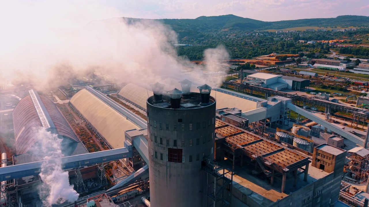 Impianto industriale con ciminiere che emettono vapore sotto la luce del sole. Una vista aerea mostra una torre di cemento che rilascia vapore bianco in un complesso manifatturiero vicino a colline.