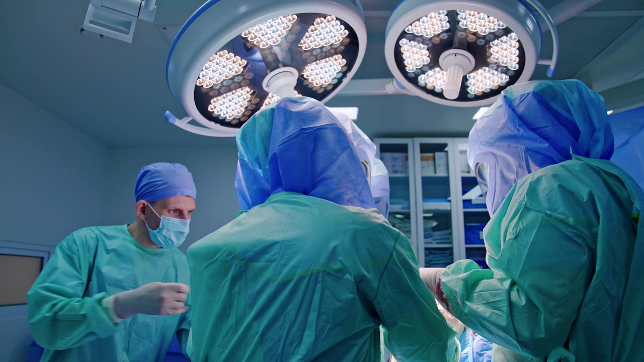 Due chirurghi in camici blu e caschi protettivi stanno di spalle alla telecamera. Un assistente maschio passa lo strumento metallico all'équipe medica.