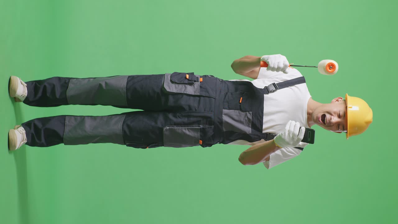 Full Body Of Asian Man Painter Wearing Safety Helmet Looking At Smartphone, Smiling, And Saying Wow While Standing In The Green Screen Background Studio