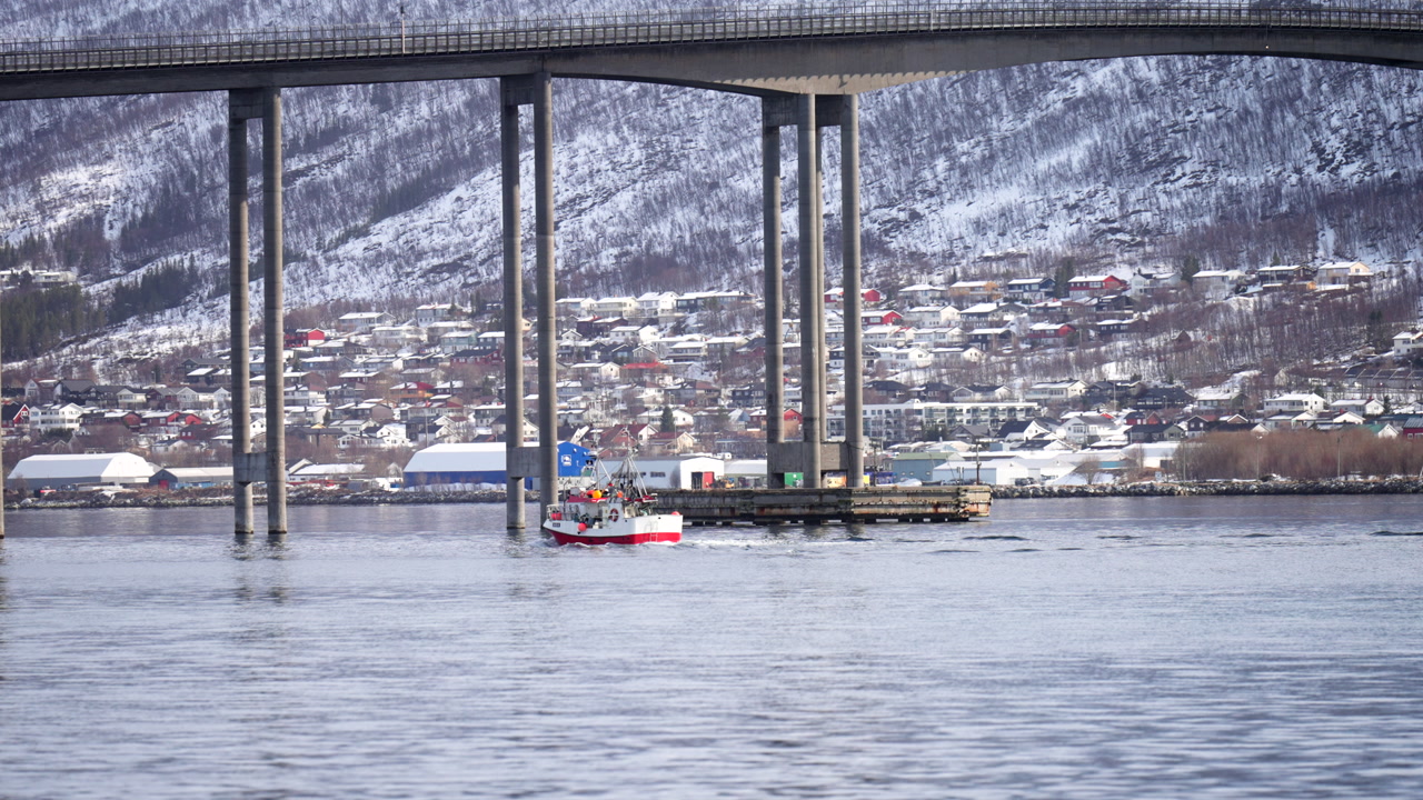 Tromsø Bridge spans across the fjord with a fishing boat below and snowy mountains in the background, capturing scenic architecture and winter life in northern Norway