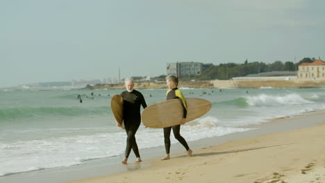 Älteres-Paar-Im-Neoprenanzug,-Das-Mit-Surfbrett-Am-Strand-Entlang-Geht-Und-Sich-An-Den-Händen-Hält