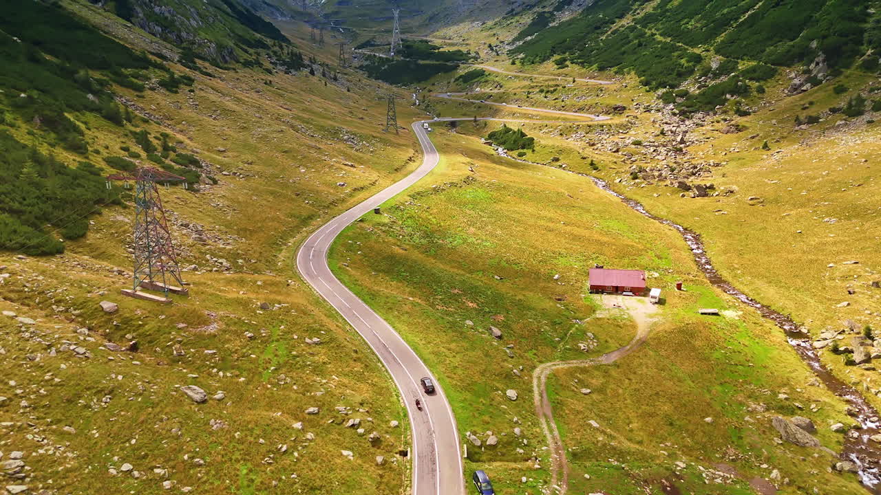 Following a car and bike riding by the wavy road in the green valley. Revealing view on the stunning Transfagarash highway and the Carpathian Mountains covered with grey clouds at the tops