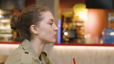 side view of a youg woman eating popcorn and drinking while talking with someone sitting at table in the cinema snack bar