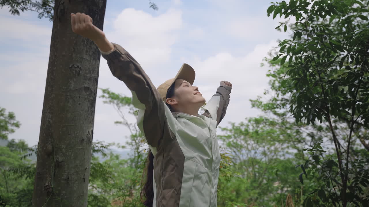 Happy woman hiker backpacker with trekking clothes reaching up top of mountain peak, raised arms hands, tourist take a deep breath in the fresh air with closed eyes surrounded lush tropical forest