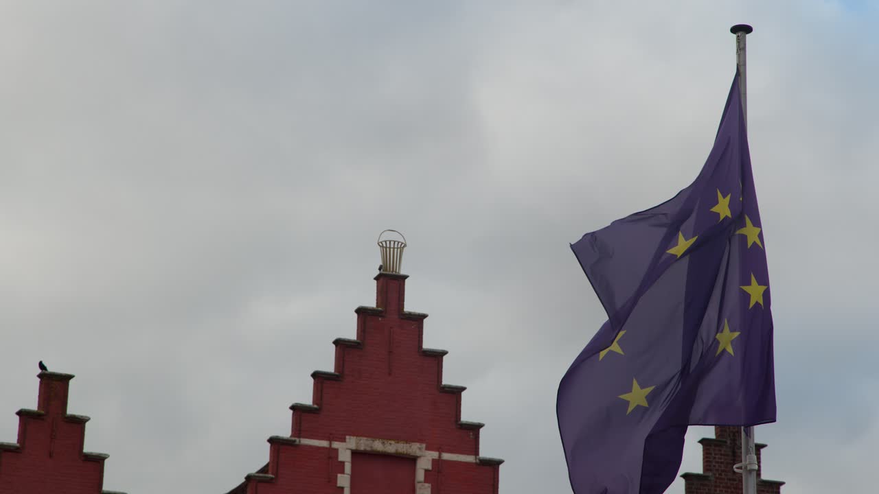 La bandiera dell'Unione Europea sventola nel forte vento sopra un edificio rosso con tetto a due falde, luce diurna nuvolosa, scatto statico
