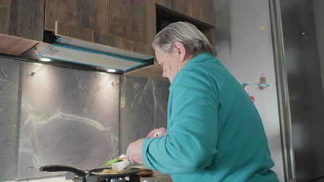 Older woman cooking in modern kitchen, stirring eggs in frying pan with spatula, focused on cooking process with kitchen tools, stovetop, and food visible in home kitchen setting