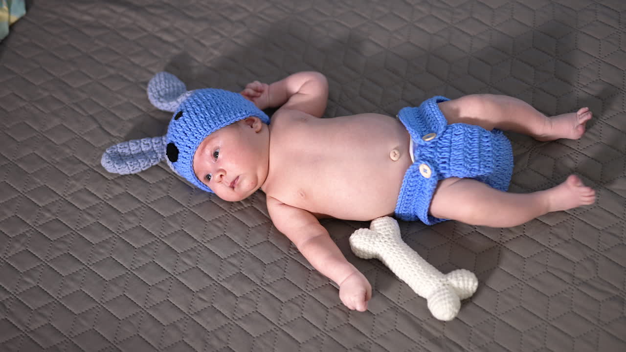 Little infant boy wearing funny hat and pants lies on the bed. Beautiful baby in puppy costume on the grey backdrop.