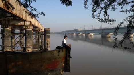 Man-Sitting-On-Steel-Deck-By-The-River-In-Mumbai,-India-Watching-The-Local-Trains-Moving-On-The-Elevated-Tracks---zoom-in-slowmo-shot