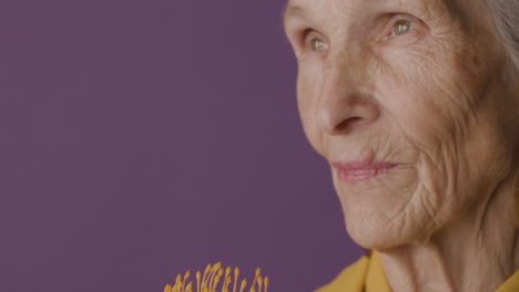 Close-Up-View-Of-Senior-Woman-With-Short-Hair-Wearing-Mustard-Colored-Shirt-And-Jacket-And-Earrings-Posing-Holding-A-Flower-On-Purple-Background