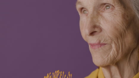 close up view of senior woman with short hair wearing mustard colored shirt and jacket and earrings posing holding a flower on purple background